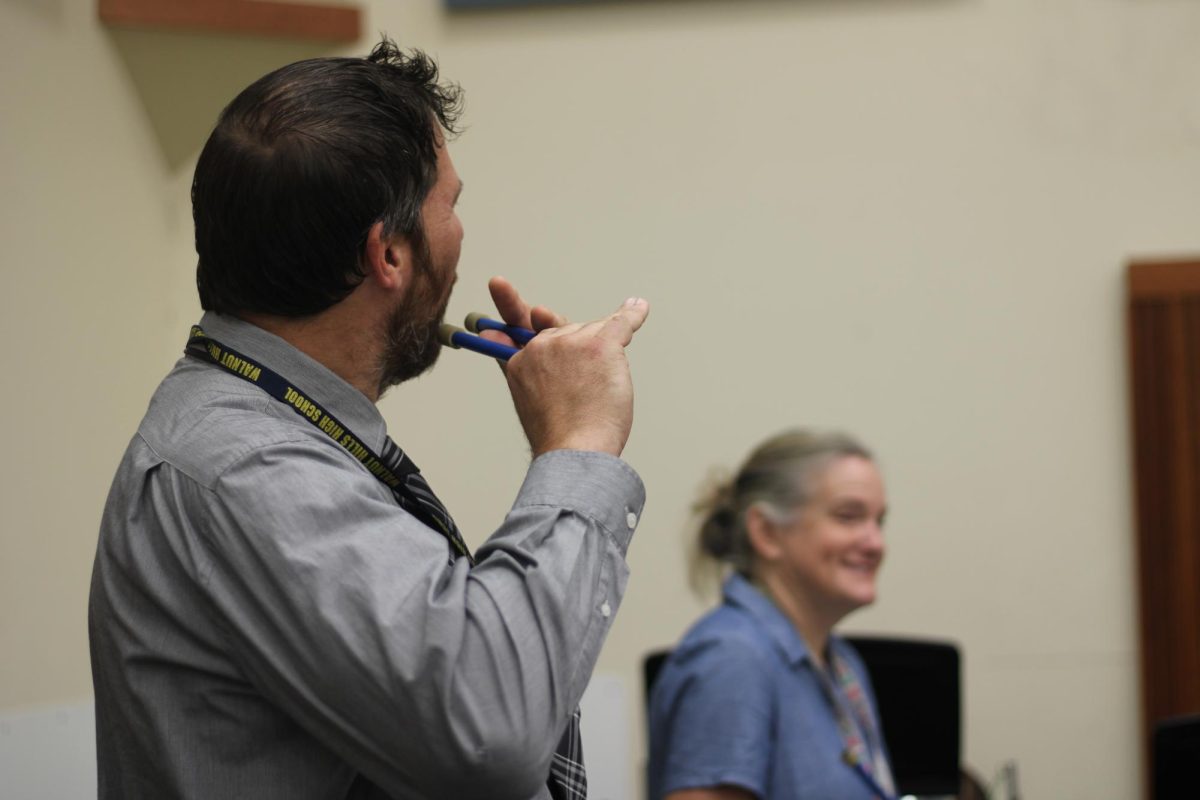 Nick Lander, Latin teacher, demonstrates his flute playing prowess while Melody Riggs, librarian, laughs in the background. "“It's difficult because the notes are not necessarily arranged like a piano," Lander said. "So we're just like low to high and you kind of have to remember where they are.”